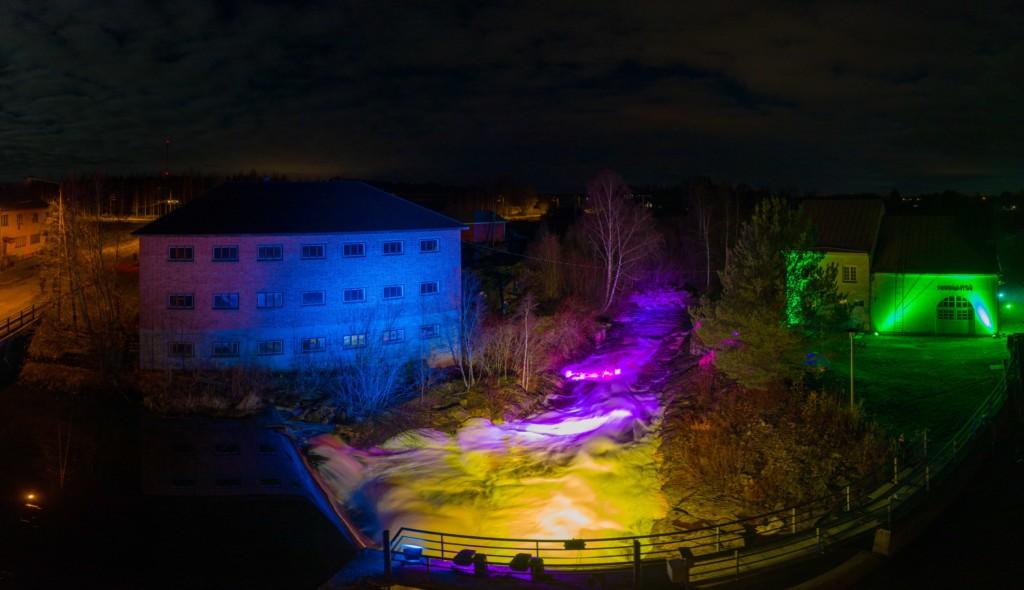 Jylhänkoski valaistuna eri värisillä valoilla. Vesi virtaa jokea pitkin rakennusten välistä