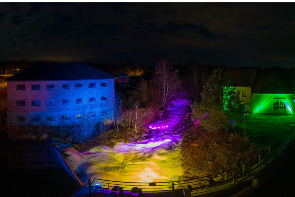 Jylhänkoski pimeällä valaistuna keltaisella ja violetilla. Viereinen voimalaitos valaistu vihreällä ja tiilirakennus sinisellä.