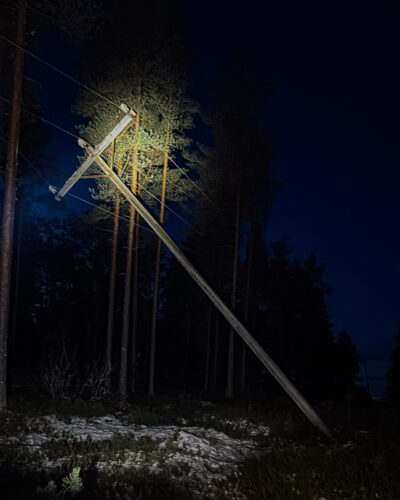 Vinossa olevaan sähköpylvääseen osuu spottivalo. Kuva on muuten pimeä. Taustalla erottuu hieman metsää ja lunta.