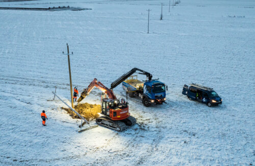 Kaksi asentajaa, kaivinkone, kuorma-auto ja pakettiauto lumisella pellolla vaihtamassa kaatunutta sähköpylvästä uuteen.